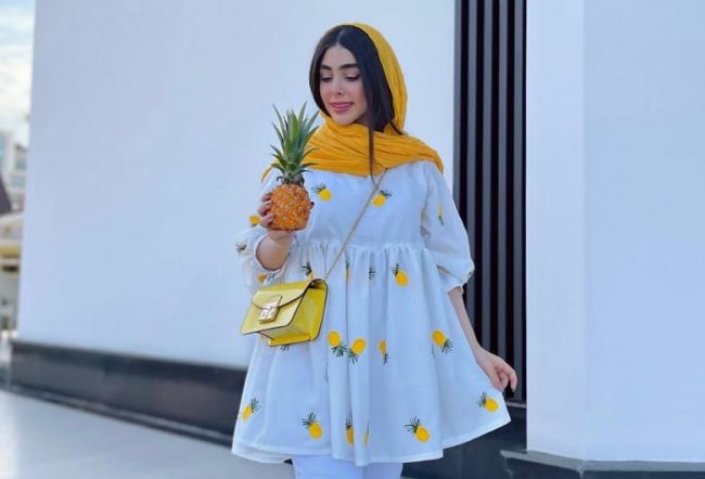 دختری با مانتو پر فروش تابستانی عروکسی که روی پارچه اش آناناس داره و یک آناناس واقهی هم در دستش گرفته است و شال سرش نیز نارنجی است